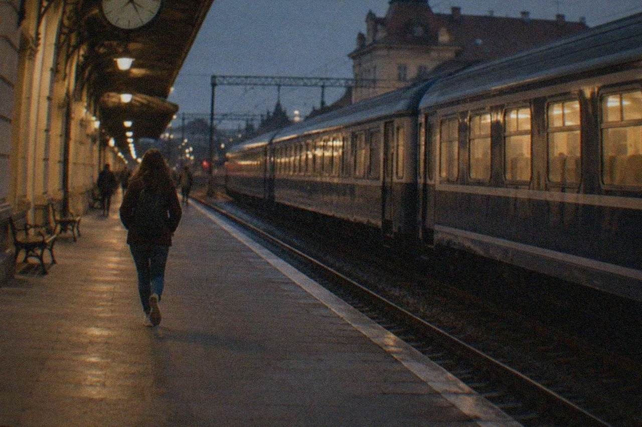 A passenger train waiting at a platform after dark, with lit carriage windows and a station clock visible