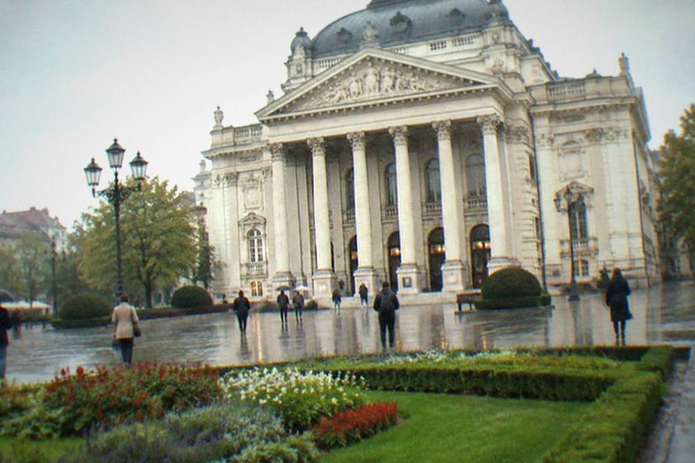 An ornate opera house facade above a wide square with flower beds and pedestrians