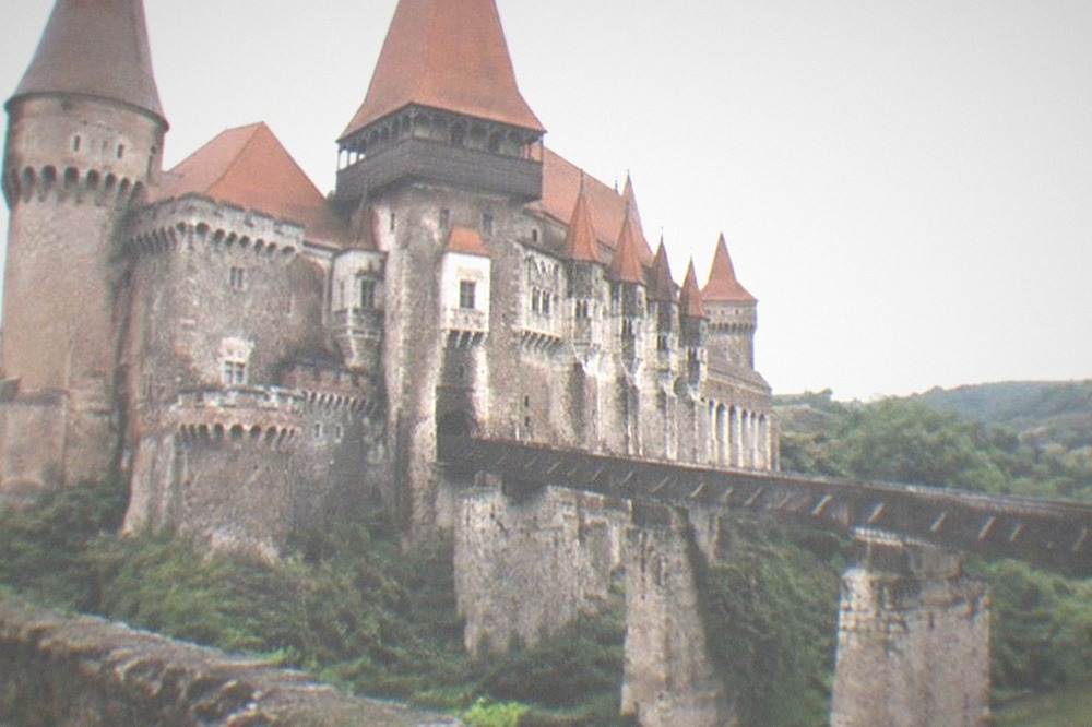 A castle tower rising above red rooftops and green hills under a summer sky