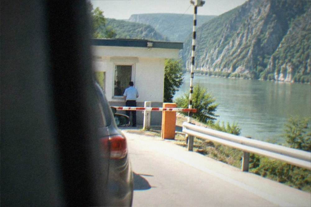 A small border post building beside a road with barriers, a striped pole and green hills behind