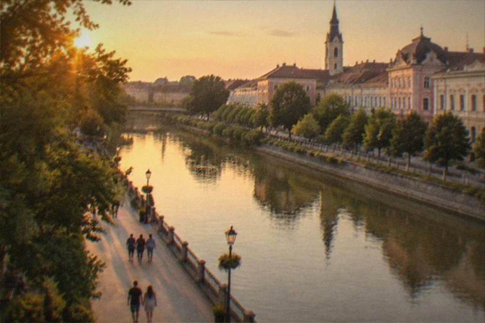 A riverside promenade in warm evening light, with people strolling and trees in full leaf