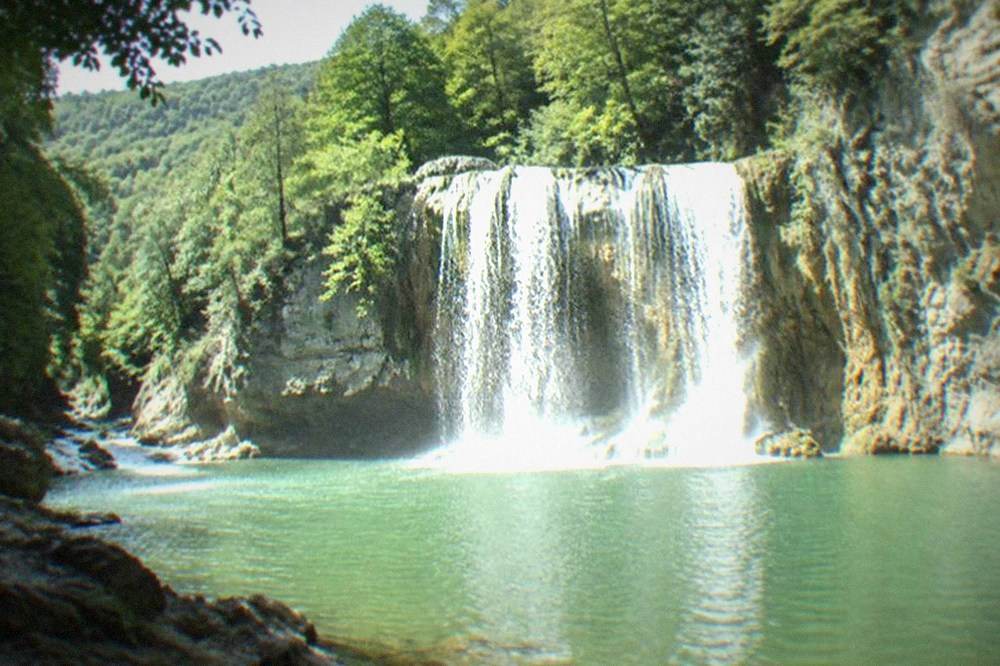 A broad waterfall pouring over a limestone ledge into a green pool in a forest