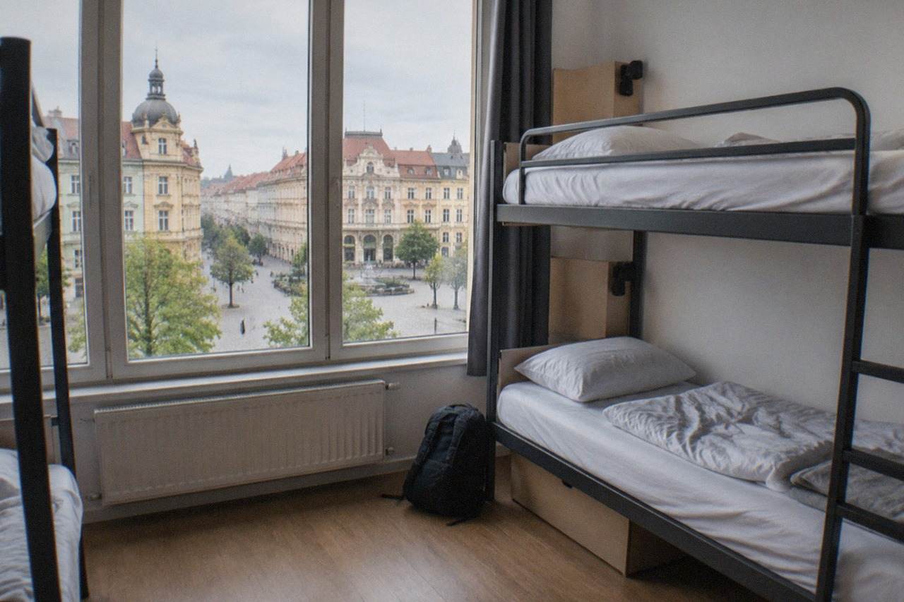 A bright hostel dorm room with bunk beds and large windows overlooking a European old town square