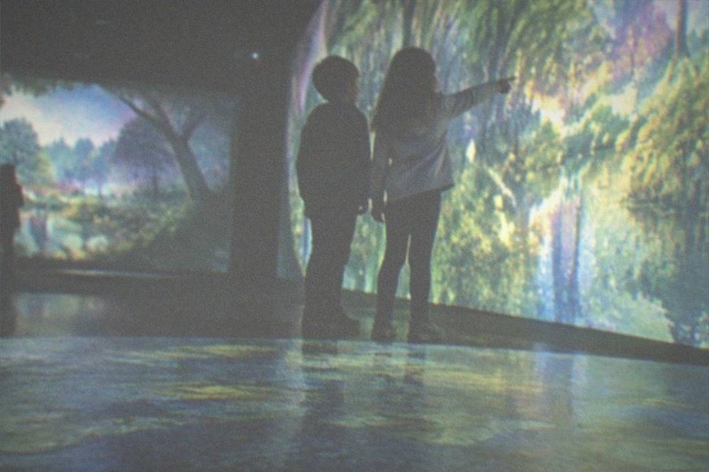 Children pointing at projected colorful artwork inside a dark immersive museum hall