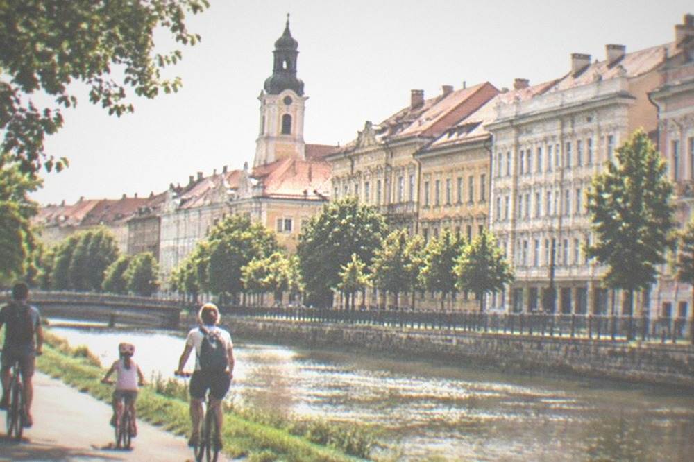 A family cycling along a green canal-side path in Timisoara with the water sparkling beside them